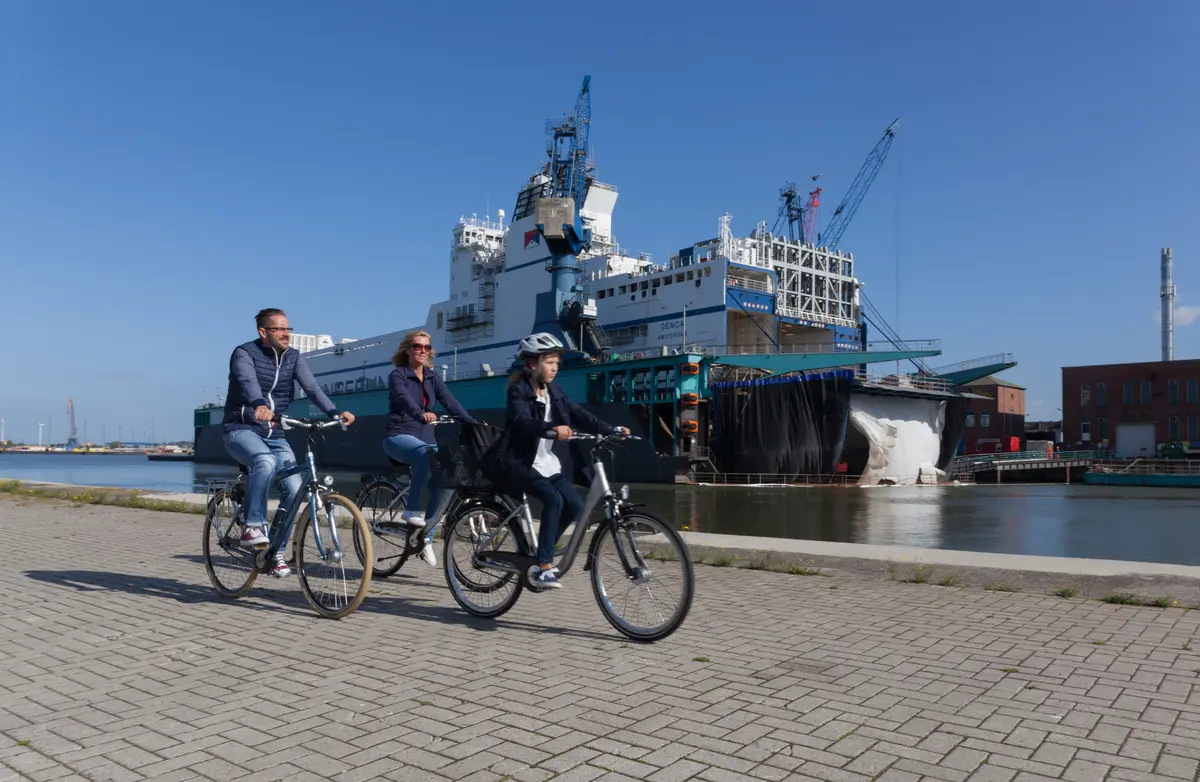 Drei Personen fahren bei Sonnenschein auf Fahrrädern an der Hafenkante in Bremerhaven entlang, im Hintergrund steht ein Frachtschiff.