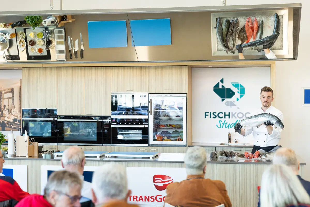 Ein Koch präsentiert bei einer Vorführung im Fischkochstudio Bremerhaven einen frischen Fisch vor Publikum.