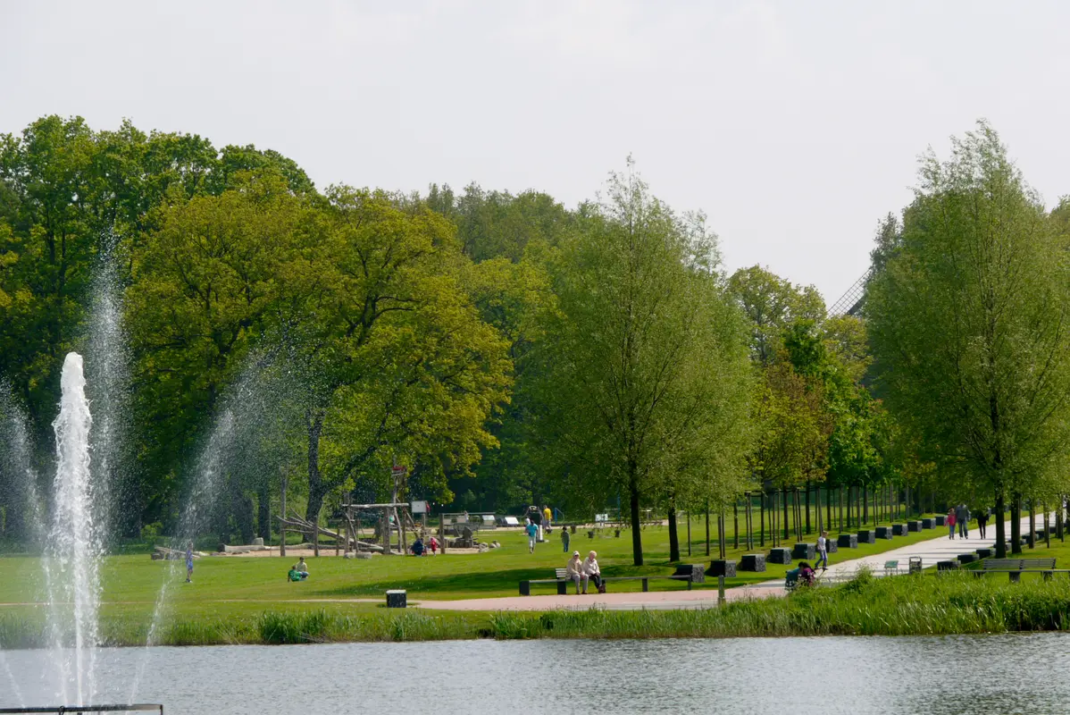Blick über einen Teich mit großer Wasserfontäne auf eine grüne Parkanlage in Bremerhaven mit Spaziergängern.