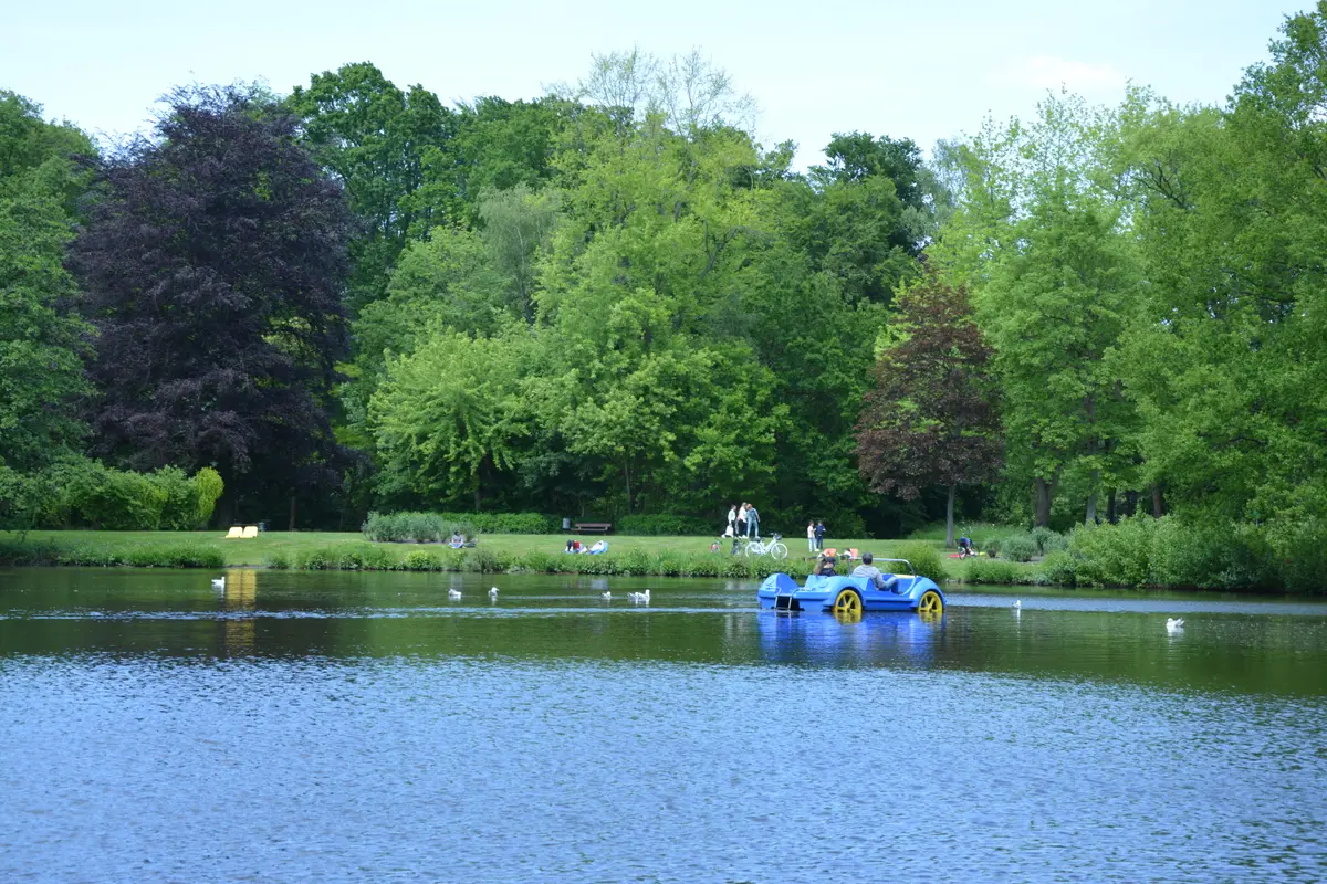Ein blaues Tretboot mit Fahrgästen fährt auf dem Parksee im Bürgerpark Bremerhaven, umgeben von Bäumen.