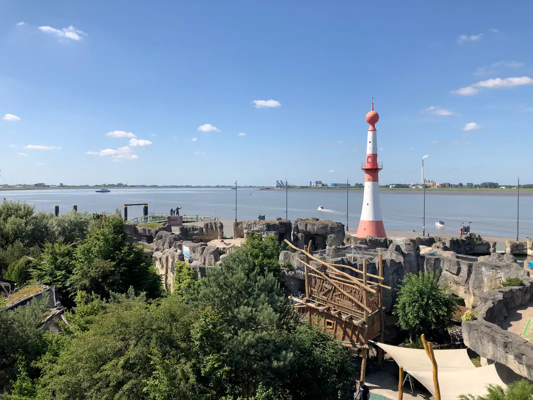 Blick über Felsgehege und Holzbauwerke im Zoo am Meer in Bremerhaven mit rot-weißem Leuchtturm an der Weser.