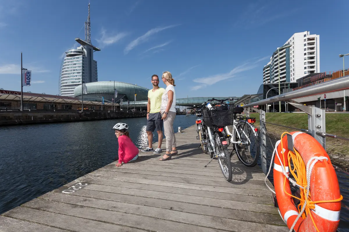 Eine Familie mit Fahrrädern macht eine Pause auf einem Holzsteg am Wasser in Bremerhaven. Im Hintergrund sind moderne Gebäude zu sehen.