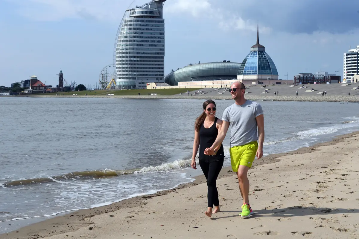 Ein Paar geht Hand in Hand am Sandstrand im Weser-Strandbad in Bremerhaven spazieren. Im Hintergrund türmt sich die Skyline auf.