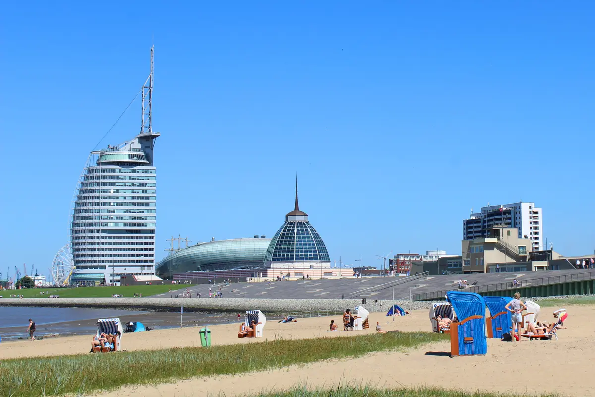 Sandstrand in Bremerhaven mit blauen Strandkörben und Menschen im Vordergrund sowie der Skyline der Havenwelten im Hintergrund.