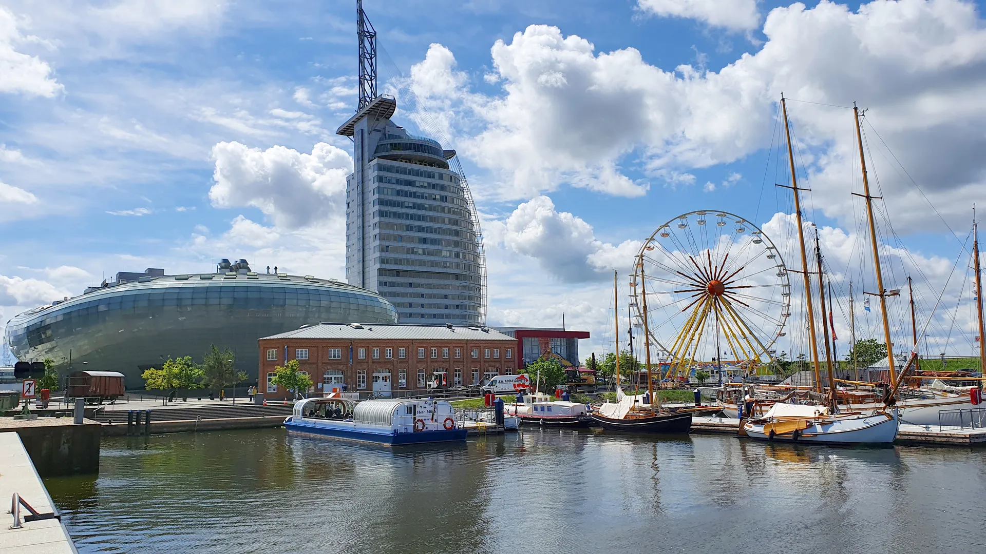 Blick über das Wasser im Hafen von Bremerhaven auf historische Segelschiffe, ein Riesenrad und prägende moderne Bauten unter bewölktem Himmel.