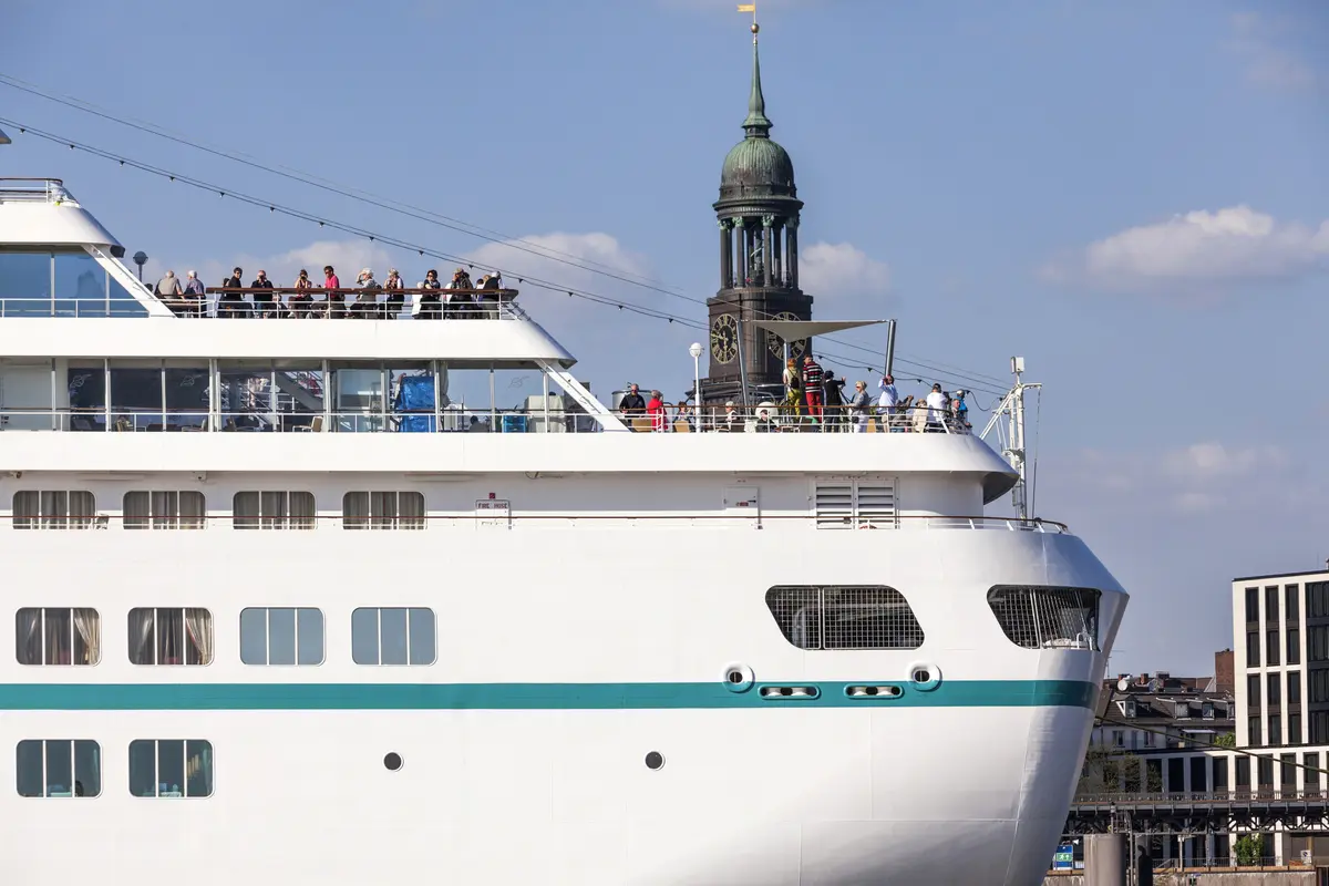 Ein weißes Kreuzfahrtschiff mit Passagieren an Deck fährt an der St. Michaelis Kirche in Hamburg vorbei.