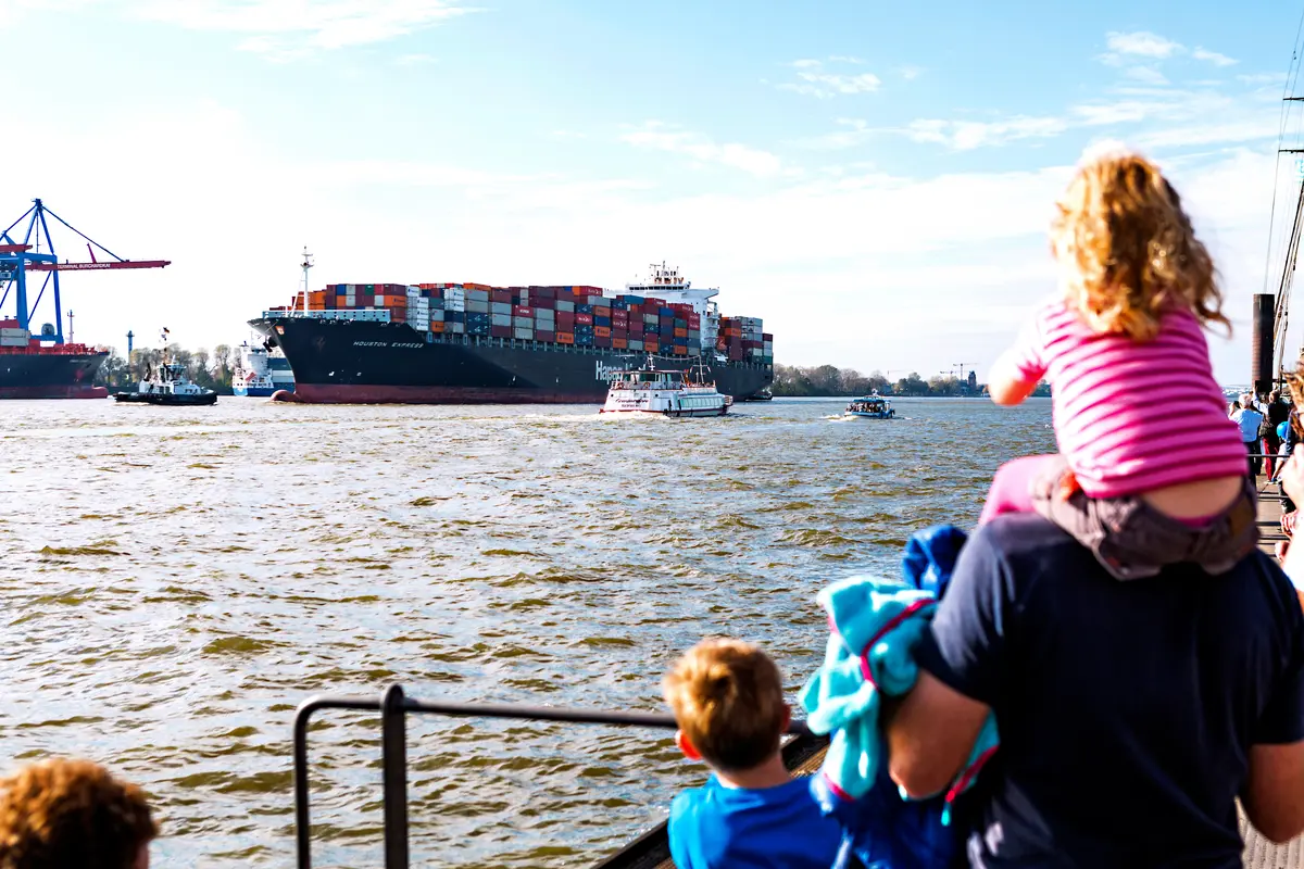 Eine Familie schaut von den Landungsbrücken in Hamburg auf die Elbe, wo ein großes Containerschiff vorbeifährt.