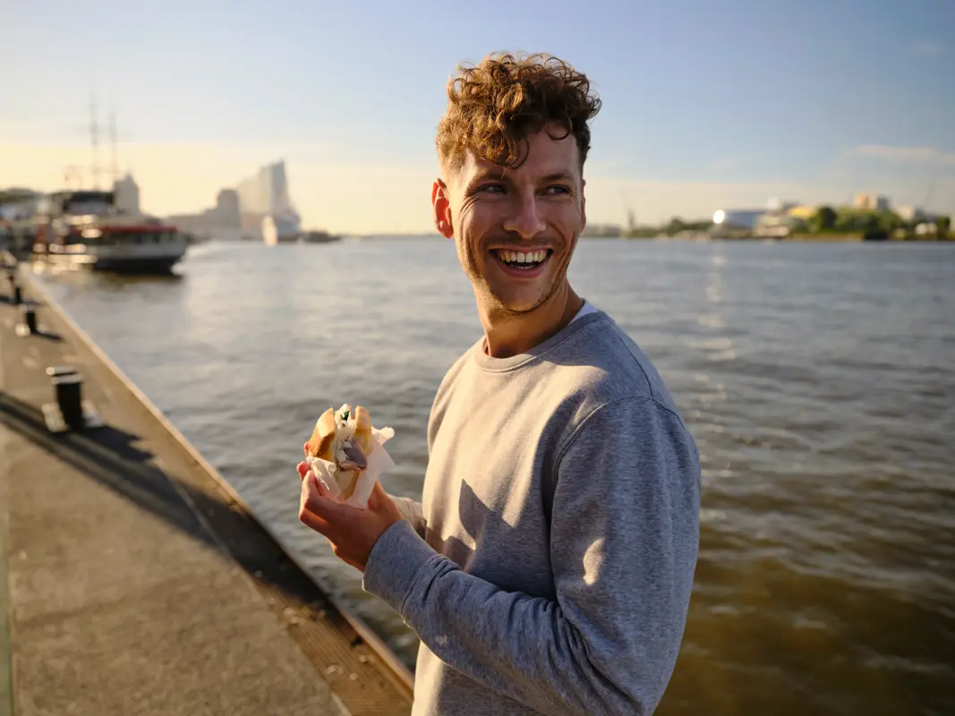 Ein lächelnder junger Mann hält ein Fischbrötchen an den Landungsbrücken im Hamburger Hafen, im Hintergrund die Elbphilharmonie.
