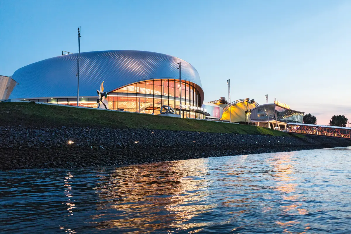 Das illuminierte Stage Theater an der Elbe in Hamburg in der Abenddämmerung mit spiegelndem Wasser im Vordergrund.