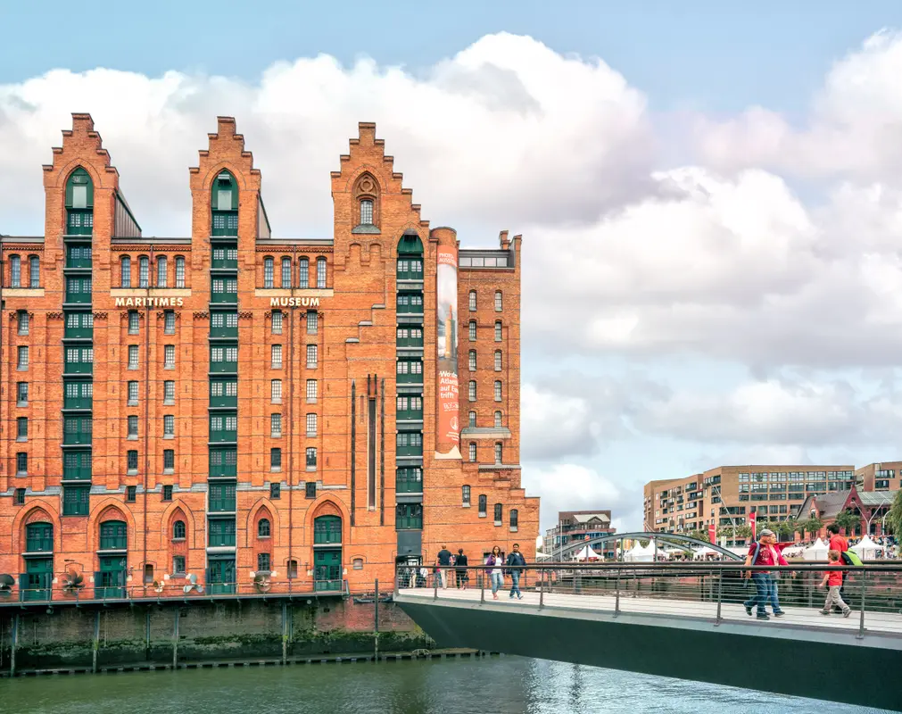 Historisches Backsteingebäude des Maritimen Museums in Hamburg an einem Gewässer mit Fußgängerbrücke und Passanten.