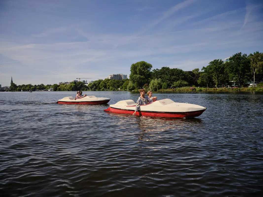 Junge Menschen fahren in rot-weißen Tretbooten über das Wasser der Alster in Hamburg unter blauem Himmel.