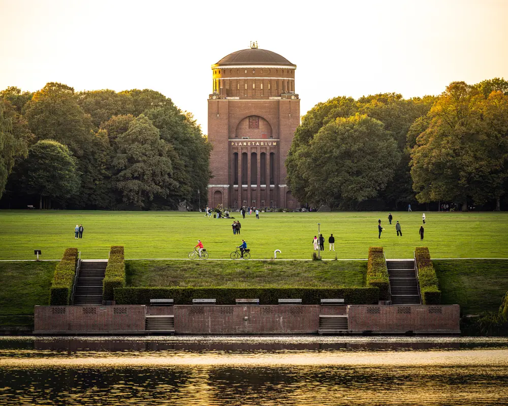 Blick auf das Planetarium im Stadtpark in Hamburg, davor eine weite Wiese mit Spaziergängern und Wasser im Vordergrund.