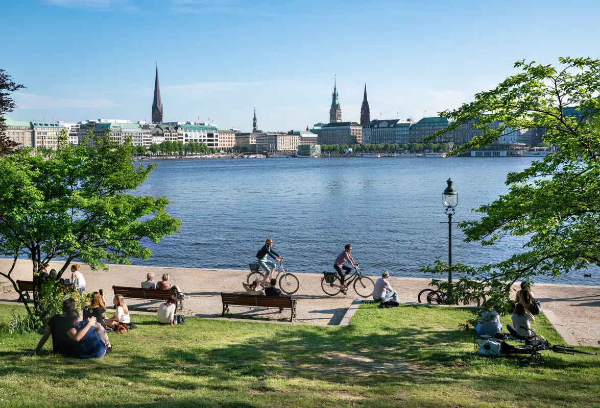Menschen entspannen im Gras und fahren Fahrrad an der Binnenalster in Hamburg vor der Kulisse der Innenstadt.
