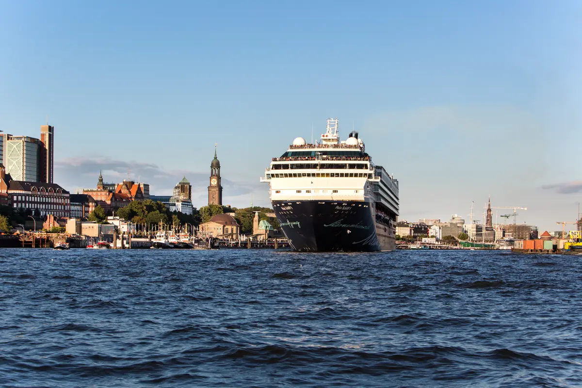 Ein großes Kreuzfahrtschiff fährt auf der blauen Elbe vor der Hafenkulisse und der Skyline von Hamburg.
