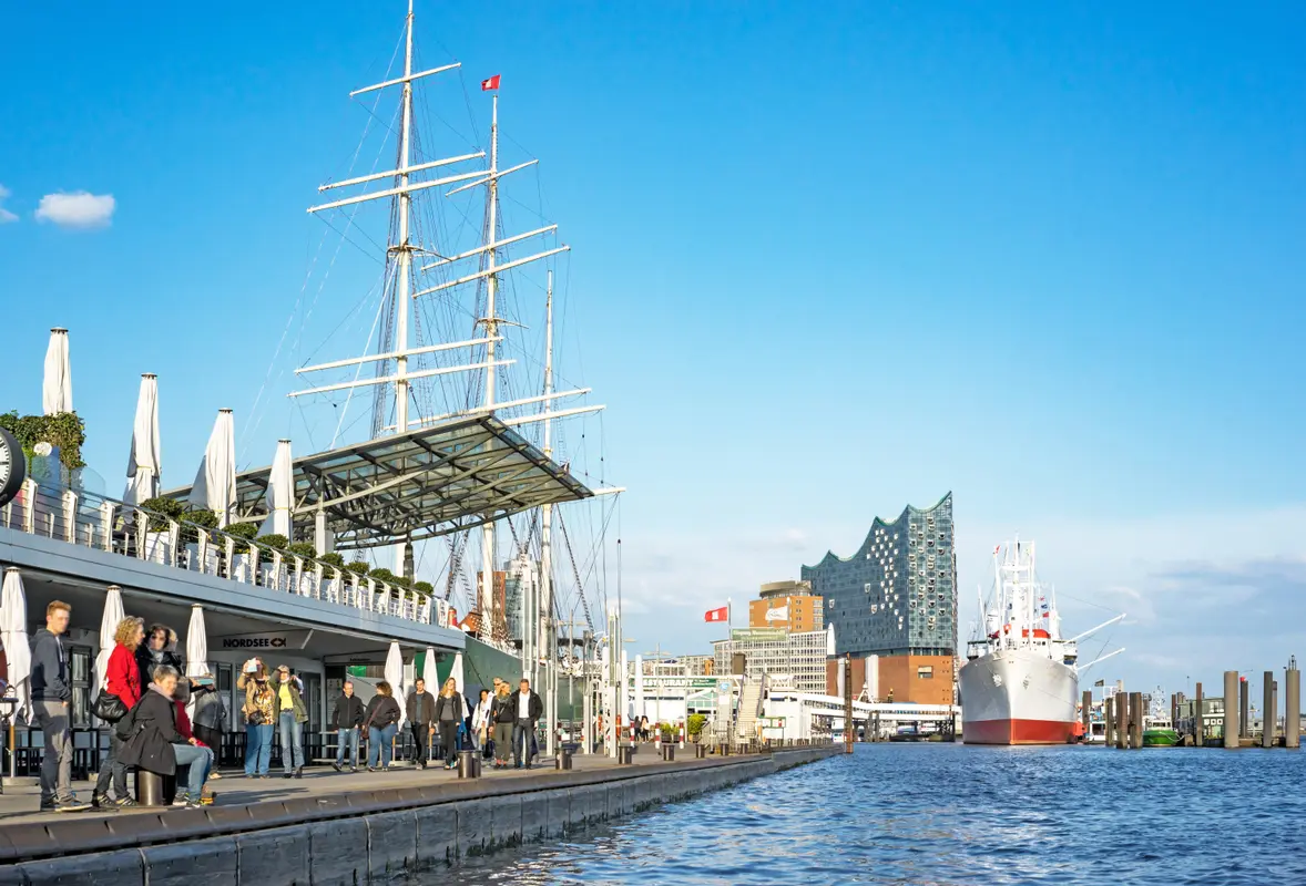 Promenade an den Hamburger Landungsbrücken mit Passanten, historischen Schiffen und der Elbphilharmonie am Wasser.