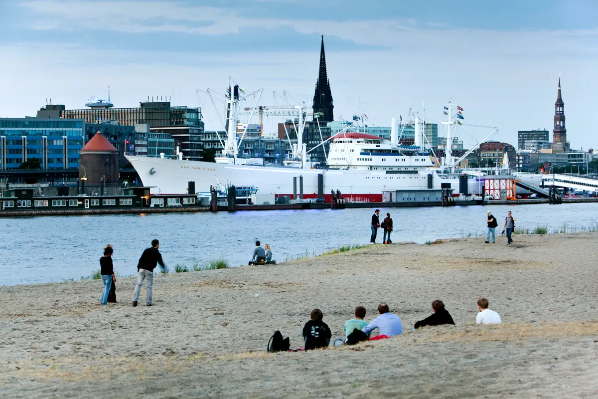 Menschen entspannen am Sandstrand in Hamburg mit Blick auf das Museumsschiff Cap San Diego und Kirchtürme.