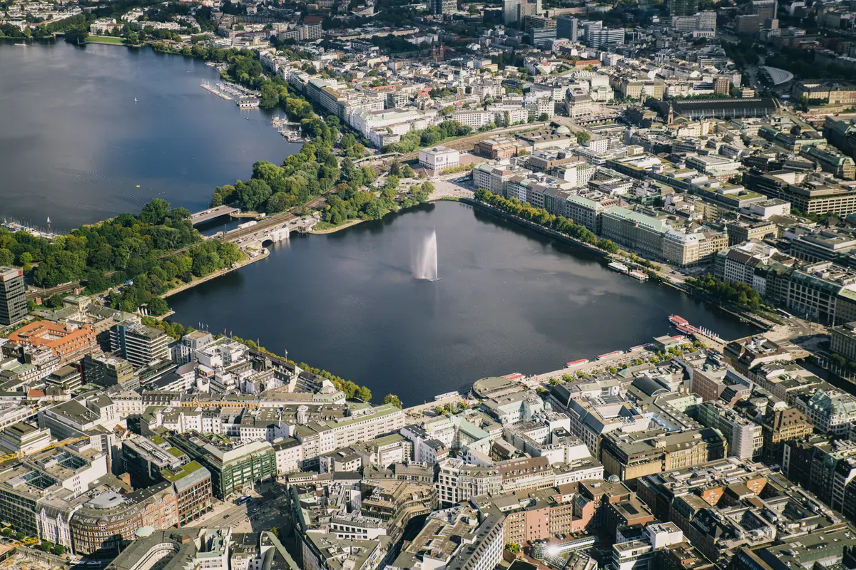 Vogelperspektive auf die Binnenalster in Hamburg mit zentraler Wasserfontäne, umgeben von Stadtgebäuden und Straßen.