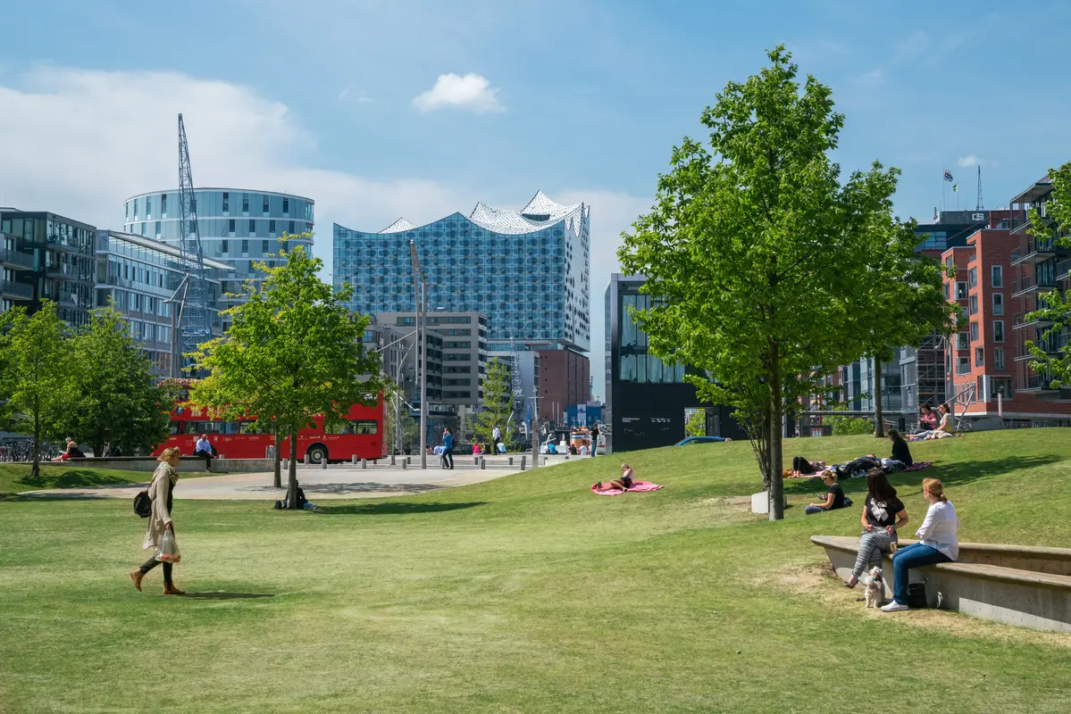Menschen entspannen auf einer grünen Rasenfläche im Sandtorpark in Hamburg, im Hintergrund ist die Elbphilharmonie zu sehen.