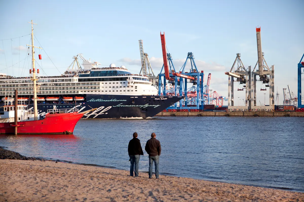 Zwei Personen stehen am Sandstrand in Hamburg und blicken auf ein großes Kreuzfahrtschiff und Hafenkräne.
