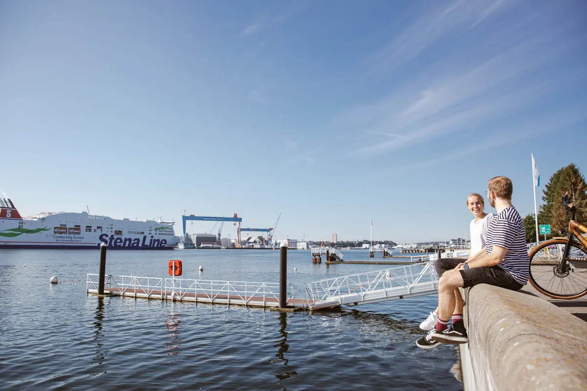 Zwei Personen sitzen entspannt auf einer Kaimauer in Kiel, im Hintergrund schwimmt eine große Fähre auf der Kieler Förde vor Werftkränen.