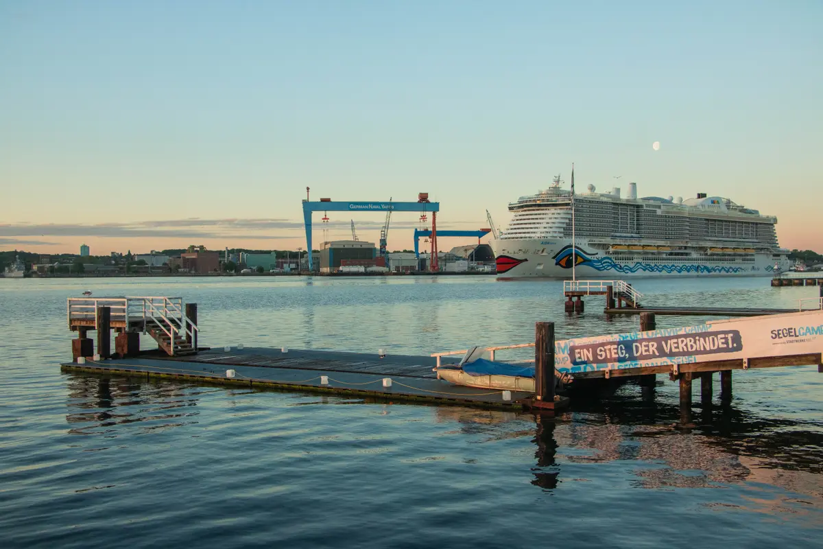 Ein Holzsteg auf dem Wasser in Kiel, im Hintergrund liegen ein Kreuzfahrtschiff und Werftkräne am Ufer.