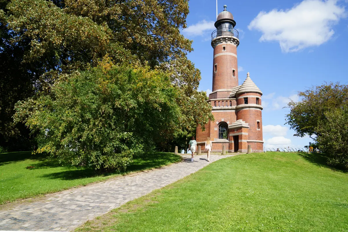 Ein Pflasterweg führt durch eine grüne Parkanlage zum historischen Leuchtturm Holtenau in Kiel.