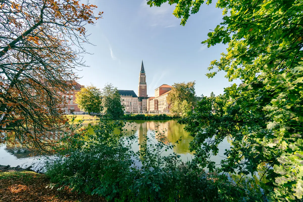 Blick durch herbstliche Baumkronen über ein Gewässer auf den Rathausturm und das Opernhaus in Kiel mit Spiegelung im Wasser.