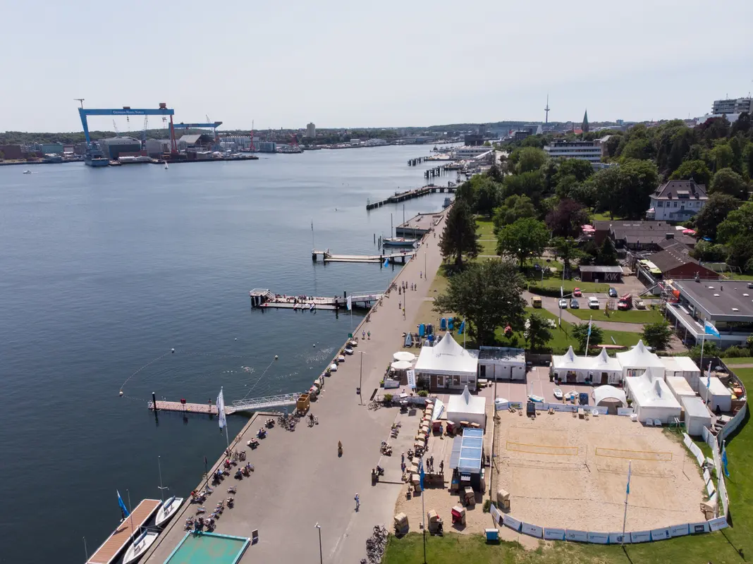 Luftaufnahme der Promenade Kiellinie in Kiel mit Beachvolleyballfeldern, weißen Zelten und Blick auf das Wasser der Förde.