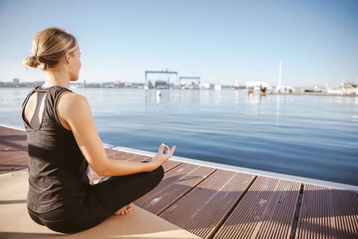 Eine Frau sitzt entspannt im Meditationssitz auf einer Yogamatte an einem Steg in Kiel mit Blick auf die Förde.
