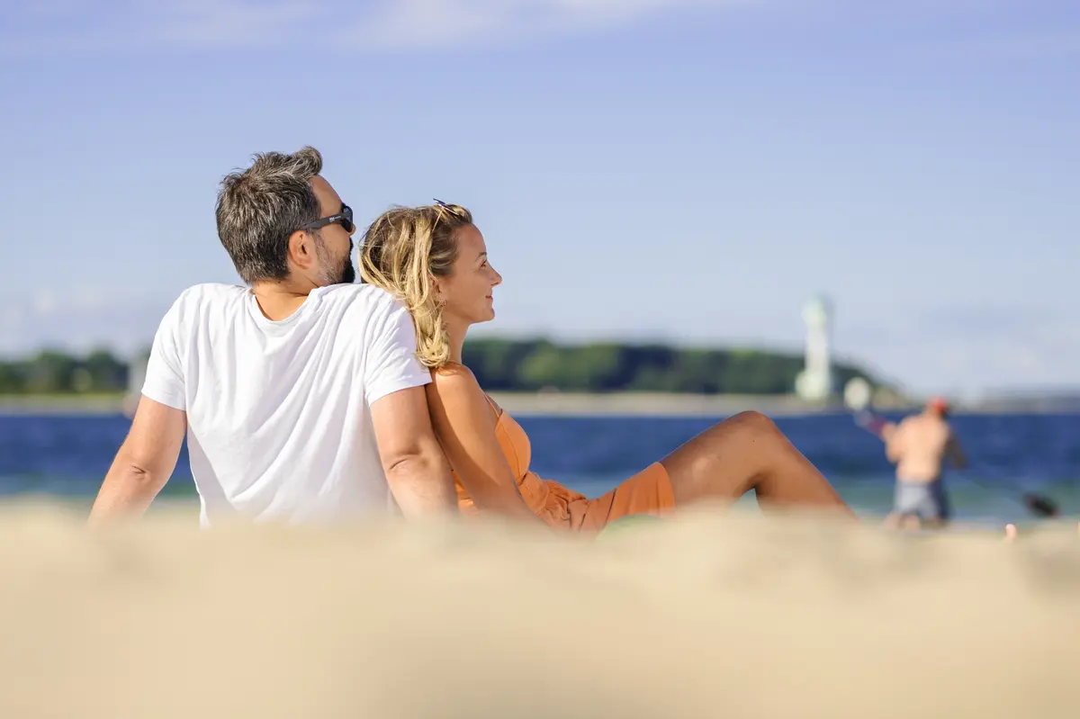 Ein Mann und eine Frau sitzen entspannt im Sand am Strand in Kiel und blicken über das blaue Meer.
