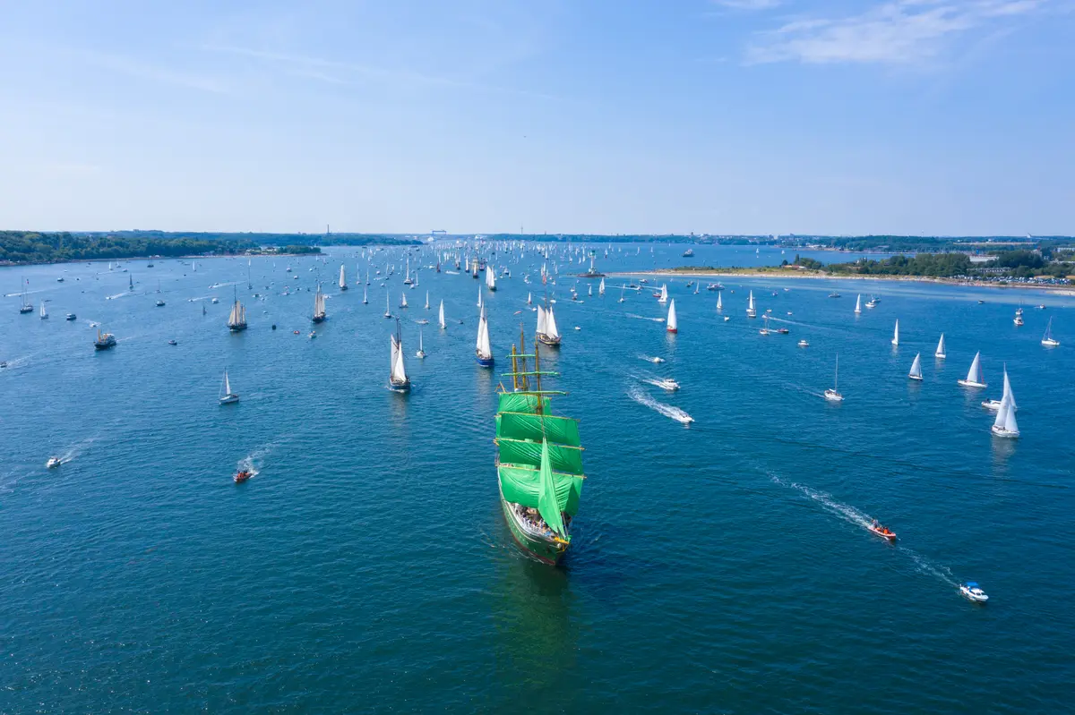 Luftaufnahme der Kieler Förde voller Segelschiffe, im Zentrum ein Großsegler mit auffällig grünen Segeln.
