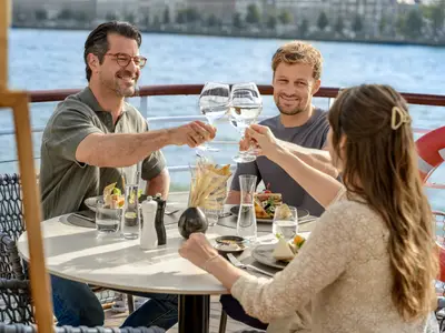 Drei lächelnde Personen toasten auf dem sonnigen Deck eines Flusskreuzfahrtschiffs mit Weingläsern, genießen Essen und den Blick auf das Wasser.