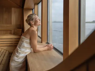 Eine Frau im Saunabereich eines Flussschiffs blickt mit geschlossenen Augen entspannt durch ein Panoramafenster auf den Fluss.