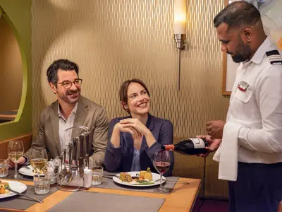 Ein lächelndes Paar sitzt im eleganten Restaurant eines Flussschiffs und beobachtet, wie ein Kellner Rotwein in ein Glas füllt.