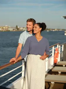 Ein diverses Paar lächelt und blickt von einem Sonnendeck eines Flusskreuzfahrtschiffs auf das Wasser und die Stadt im Hintergrund.