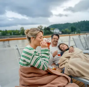 Drei Personen auf einem Flussschiff-Sonnendeck, in Decken gekuschelt. Eine Frau trinkt, eine andere schläft, ein Mann lächelt. Bewölkte Flusslandschaft mit Hügeln.