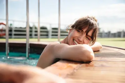 Lächelnde Frau im Pool auf einem Flusskreuzfahrtschiff, die sich auf den hölzernen Poolrand lehnt. Heller Taghimmel im Hintergrund.