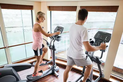 Ein Paar trainiert in einem hellen Fitnessraum an Bord eines Schiffes auf Crosstrainern mit Blick auf das Wasser.