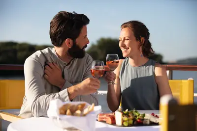 Ein Paar lacht und stößt mit Roséwein auf dem Sonnendeck eines Flussschiffs an, mit Käse und Brot auf dem Tisch.