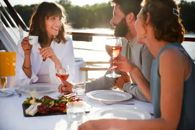 Drei Freunde genießen Wein und eine Antipasti-Platte an einem Tisch im Freien auf einem sonnigen Schiffsdeck.