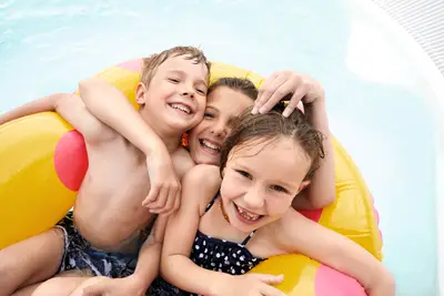 Drei fröhliche Kinder sitzen in einem gelb-roten Schwimmring im Pool und lachen in die Kamera.