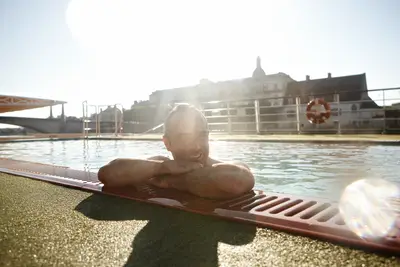 Lächelnder Mann entspannt im Pool auf dem Sonnendeck eines Flussschiffes, mit sonniger Stadtkulisse im Hintergrund.