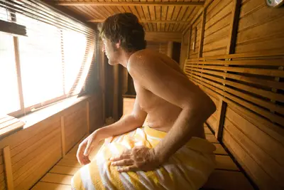 Ein Mann mit Handtuch entspannt in einer modernen Holzsauna, blickt aus einem großen Fenster mit Jalousien ins helle Licht.