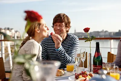 Ein lächelndes Paar frühstückt auf dem Sonnendeck eines Flussschiffs, der Mann füttert die Frau liebevoll mit einer Erdbeere. Eine rote Rose schmückt den Tisch.
