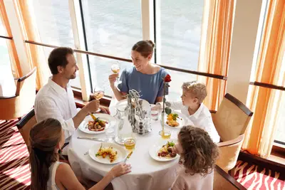 Eine Familie mit zwei Erwachsenen und drei Kindern genießt eine Mahlzeit an einem runden Tisch in einem hellen Restaurant mit Blick auf das Wasser.