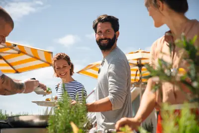 Gruppe von Menschen bei einem Outdoor-BBQ auf dem Sonnendeck. Ein Mann lächelt in die Kamera, während eine Frau Essen serviert bekommt.