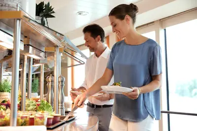 Ein Mann und eine Frau bedienen sich an einem eleganten Buffet mit frischen Speisen, beleuchtet von Tageslicht durch große Fenster.