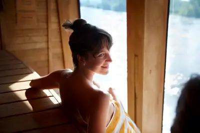 Junge Frau entspannt in einer holzverkleideten Sauna, blickt lächelnd aus dem Fenster auf ruhiges Wasser. Sie trägt ein gestreiftes Handtuch.