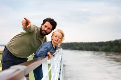 Vater und Sohn auf einem Flusskreuzfahrtschiff, der Vater zeigt über die Reling auf den Fluss, der Sohn lächelt in die Kamera.