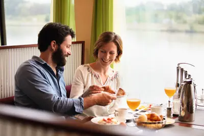 Lächelndes Paar beim Frühstück auf einem Flussschiff, teilt Croissants, mit Orangensaft und Kaffee vor Flusslandschaft.